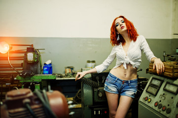 Red haired girl wear on short denim shorts and white blouse with steel spring at hand posed at industrial machine at the factory.