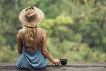 Rural woman with cup of coffee siting and look on jungle forest. Back view.
