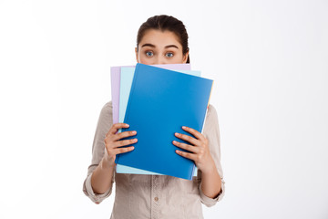 Surprised young beautiful brunette businesswoman holding colorful folders over white background