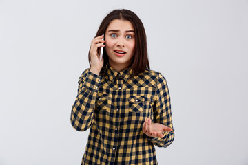 Upset young beautiful girl dressed in plaid shirt speaking on phone, looking at camera over white...