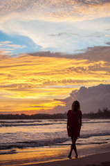 Fototapeta premium Romantic blond girl in red dress walking on the ocean shore during sunset.