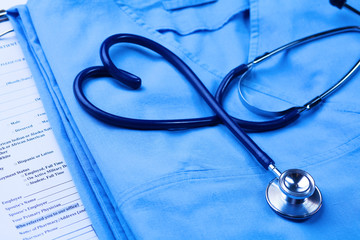 Medical stethoscope twisted in heart shape lying on patient medical history list and blue doctor uniform closeup. Medical help or insurance concept. Cardiology care, health, protection and prevention