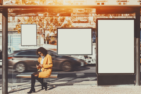 Curly Brunette Woman In Sunglasses And Yellow Coat Working On Her Digital Tablet While Sitting And Waiting Bus Inside Of Glass City Bus Stop, With Several Blank Mock-up Banners Around Her