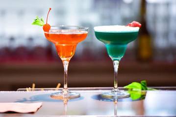 Two cocktails on the bar counter. One cocktail is green and the other is orange. They are decorated with fruits ( cherry and raspberry ). Bottles in blurred background