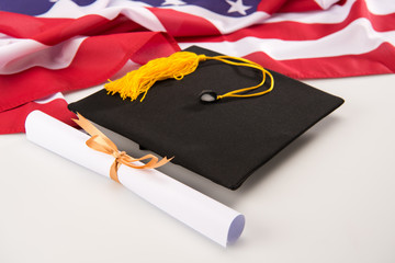 Close-up view of graduation mortarboard, diploma and us flag on grey, education concept