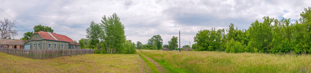 Rural landscape panorama with old wooden house behind picket and dirt road against cloudy sky background. Bolshaya Doroga village, Tambovsky region, Russia.   © shujaa_777