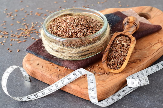 A glass bowl of flax seeds with olive scoop surrounded by artificial flowers on a grey abstract background. Healthy eating concept.