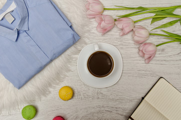 Coffee Mug, blue shirt, pink tulips, macaron cake and notebook with blank pages on the table. Working concept. Top view