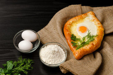 Georgian cuisine. Khachapuri on sackcloth, flour, eggs and parsley on black table, closeup