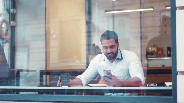 Distant view of a young businessman sitting at a table in a cafe using iphone and laughing, watching the passersby. Successful people, daily routine. Entertaining, relaxing, communicating.