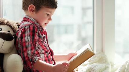child reading book, smart kid wearing checkered red shirt sitting on window and holding book in orange cover, preschooler boy reading stories on window with his fur toy friend, kid turning book pages