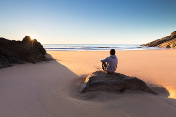 A person sits on a beautiful remote beach in Australia and watches a crisp sunrise over the ocean. - Powered by Adobe