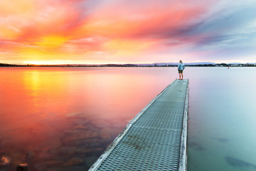 A person watches a beautiful, colourful sunset over the sea from the end of a small jetty.