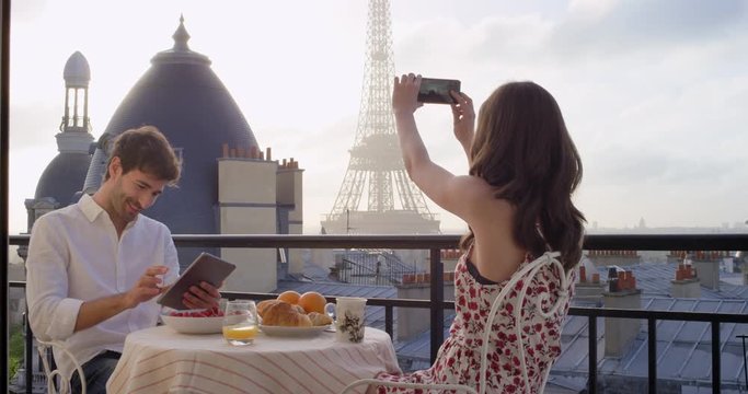Young tourist couple in Paris hotel enjoying breakfast on terrace woman photographing view of Eiffel Tower at Sunrise with smartphone in the morning