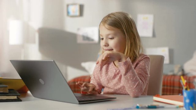 Cute Young Girl Uses Laptop Computer with Interest while Sitting at the Table in Her Light Room. Shot on RED EPIC-W 8K Helium Cinema Camera.