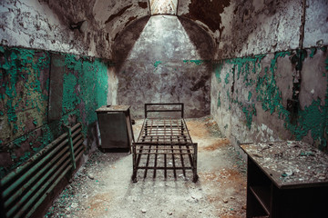 Abandoned prison cell room with old rusty bed frame and peeling walls.