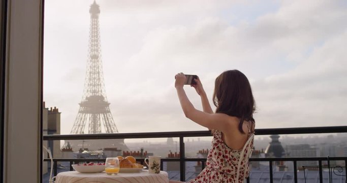 Woman taking photograph of Eiffel Tower using smartphone from hotel balcony at sunrise photographing scenic Paris cityscape background view enjoying European honeymoon vacation travel adventure