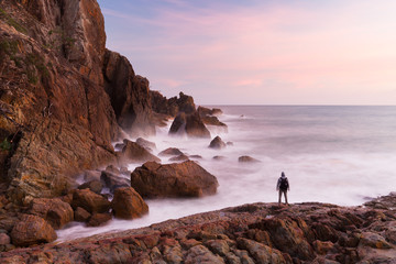 A person is silhouetted by a pink sunrise at the base of a coastal cliff in a beautiful seascape...