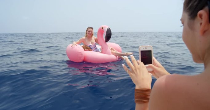 Woman lying on pink inflatable flamingo bet friend taking photograph with smartphone digital camera floating alone in middle of ocean sharing images with mobile phone technology