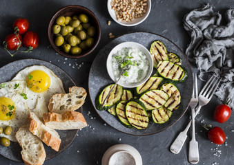 Grilled zucchini, fried eggs, olives, tomatoes, ciabatta - simple snack or appetizer. Mediterranean style food. On a dark background, top view. Flat lay