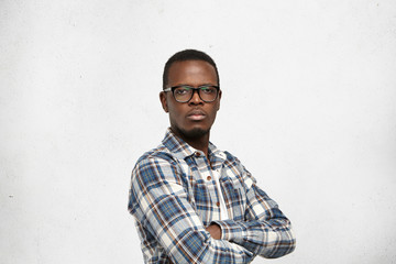 Arrogant Afro-American young hipster wearing glasses in black frame and checkered shirt looking at camera with indifferent stony facial expression, keeping his arms folded against white studio wall