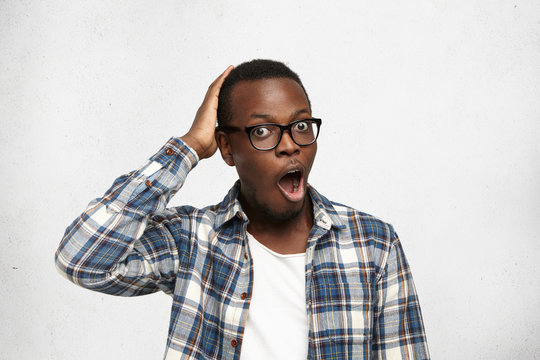 You gotta be kidding me. Close up studio shot of handsome dark-skinned man wearing eyeglasses scratching back of his head, opening mouth widely, looking at camera in surprise and full disbelief