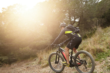 Rear shot of European professional rider in helmet, cycling clothes and eyeglasses riding black electric bike down trail with sun shining ahead of him. People, active healthy lifestyle and extreme