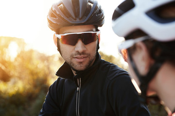 Handsome young Caucasian man with stubble wearing sunglasses and helmet having conversation with unrecognizable woman in protective gear, giving her instructions before ride. Selective focus