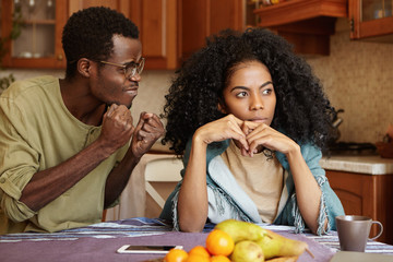 Black full of anger husband clenching fists mad at his indifferent wife, desiring for explanations, trying hard to hold himself together. African couple having serious quarrel at kitchen table