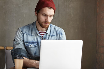 Serious handsome young European freelancer dressed in trendy clothing working remotely on laptop computer, having worried look, trying hard to finish his work in time to avoid deadline stress