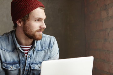 Side view of handsome stylish man sitting in front of open laptop and looking through window, watching people rushing and hurrying outside while he surfing internet at cafe, waiting for his lunch