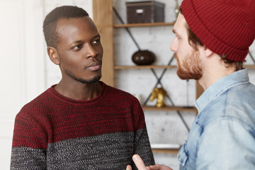 Interracial friendship, people, youth and happiness. Stylish bearded hipster in hat explaining something while having conversation or dispute with his African American friend dressed in sweater