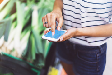 Young woman shopping healthy food in supermarket blur background. Female hands buy products and using mobile smart phone in store. Hipster at grocery holding basket. Person comparing price of produce