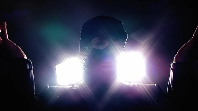 A terrorist with a balaclava covering his face, questioning and menacing us (victim's point-of-view), grabbing us with his hands. Strong lights behind him. Close-up shot.
