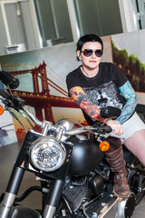 Naklejka na ściany i meble Cool sensual biker girl sitting on old fashioned motorcycle in garage interior on grey wooden wall background, vertical picture