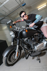 Naklejka na ściany i meble Cool sensual biker girl sitting on old fashioned motorcycle in garage interior on grey wooden wall background, vertical picture
