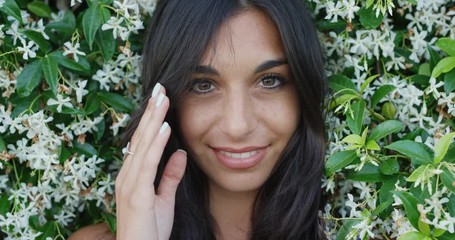 Close up portrait of beautiful young woman running hand through hair smiling in front of wall of flowers outdoors slow motion - Powered by Adobe