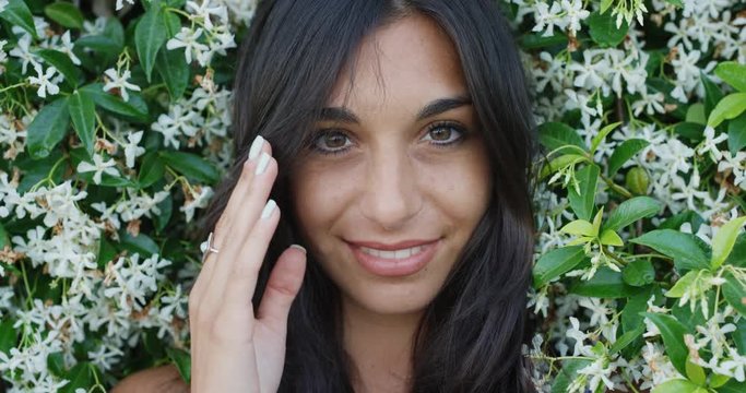 Close up portrait of beautiful young woman running hand through hair smiling in front of wall of flowers outdoors slow motion