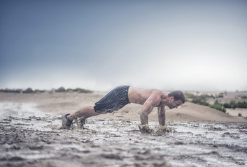 A powerful strong athletic male does a flying pushup in a muddy puddle with a rough terrain with a city in the background