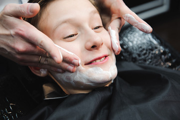 Funny boy in a black salon cape in the barbershop. Barber applies shaving foam with the help of the shaving brush on his face.