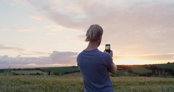 Rear view of Man taking panorama photographing wheat field using smartphone capturing epic nature background Cornwall, UK