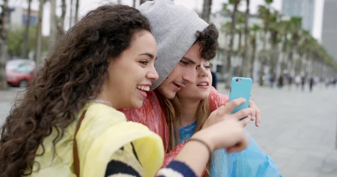Diverse group of teenage friends holding smartphone sending text snapchat message wearing multi colored raincoat poncho in rainy weather on the beach celebrating adventure 