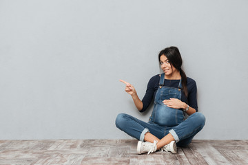 Happy pregnant lady sitting on floor pointing to copyspace