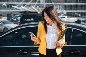 Cheerful young businesswoman holding folder and using cell phone outdoors