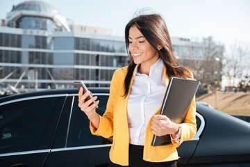 Fototapeta premium Smiling young businesswoman with documents in folder using smartphone outdoors
