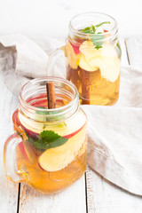 Glass mugs of tea with ice, mint, apples and cinnamon sticks top view on a white wooden background.