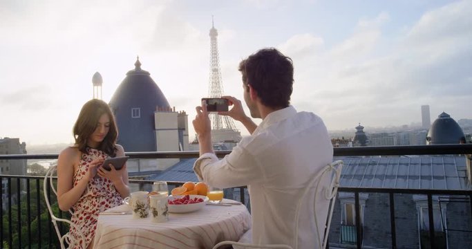 Young tourist couple in Paris hotel enjoying breakfast on terrace Man photographing view of Eiffel Tower at Sunrise with smartphone in the morning