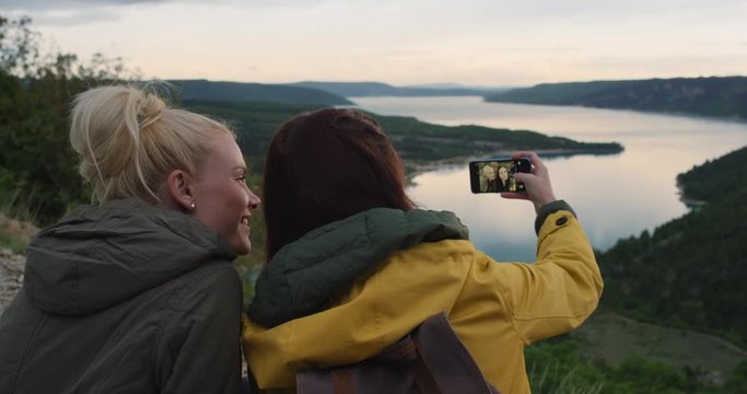 Two backpacker girl friends taking photograph of lake using smartphone women photographing scenic landscape nature background view enjoying vacation travel adventure