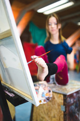 Disabled beautiful young artist painting incredible scenes in attic by holding paintbrush in her toes. Selective focus.