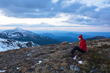 Fototapeta premium A tourist girl sits on a big gray stone and looks dreamily at mountains.